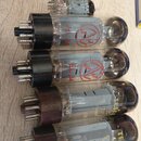 Fotografie zobrazuje skupinu elektronických vakuových elektronek různých typů a velikostí. Dvě horní větší elektronky mají černé patky a červený nápi…