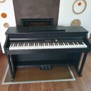 Černý digitální piano s klaviaturou a dřevěným stojanem. Na pravé straně ovládacího panelu je bílý nápis „Rhodes“. Nástroj obsahuje tři pedály a na h…