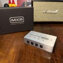 Stříbrný kovový napájecí adaptér s nápisy MXR dc brick. Na horní straně jsou označení výstupů V6, V6, V9, V9 a 3V. Vedle zařízení je černá krabice s…