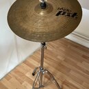 Zlatý činel z kovu s viditelným rýhováním, umístěný na chromovaném stojanu. Na povrchu je černý nápis „paiste PXT 5 MEDIUM CRASH“. Povrch činelu vyka…