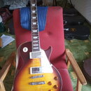 Elektrická kytara s trupem v barvě sunburst a bílým pickguardem. Na hlavě hmatníku je viditelné logo Gibson. Nástroj má dva humbuckery, čtyři zlaté o…