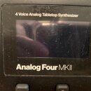 Detailní pohled na displej a ovládací panel černého syntetizátoru. Nad displejem je bílý nápis 4 Voice Analog Tabletop Synthesizer a pod ním nápis An…