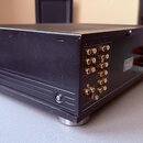 Černý audio zesilovač v kovovém šasi s žebrovaným zadním panelem a stříbrnými nožičkami. Na zadní straně jsou zlaté RCA konektory s označením L a R.…