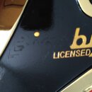 Detailní záběr na černý lesklý povrch s kovovými zlatými prvky. Na povrchu je zlatým písmem nápis „by LICENSED“. V levé části jsou vidět drobné škráb…