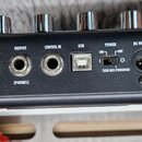 Zadní strana černého elektronického zařízení s řadou konektorů a ovládacími knoflíky nahoře. Jsou zde nápisy INPUT, OUTPUT [PHONES], CONTROL IN, USB…