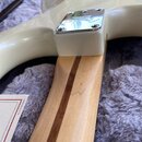 Detail bílé elektrické kytary s krkem z jasného dřeva a tmavým středovým pruhem. Na kovovém krytu sáhlého spoje je viditelné logo Fender. Vedle krku…