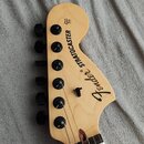 Fotografie zachycuje hlavu elektrické kytary z přírodního světlého dřeva se šesti černými ladičkami. Na povrchu je černý nápis Fender® STRATOCASTER a…