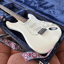 Bílá elektrická kytara typu Stratocaster s bílým pickguardem a třemi snímači leží v otevřeném černém kufru s modrým polstrováním. Na těle kytary je v…