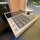 Bíle provedený sampler s mřížkou 16 šedých padů a displejem v horní části. Na spodní hraně je červený nápis AKAI a vedle něj nečitelný text. Zařízení…