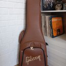 Hnědý polstrovaný pouzdro na kytaru z materiálu imitujícího kůži. V dolní části je umístěno zlaté logo „Gibson“. Pouzdro má v horní části zip s kovov…