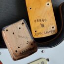 Fotografie zachycuje detaily elektrické kytary. Na zadní straně dřevěné hlavy jsou nápisy 68940 a JOHN MAYER, vedle nich je viditelné logo s obrázkem…
