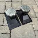 Dva černé plastové stojany s kulatými kovovými horními deskami. Stojany mají čtvercovou základnu a jsou umístěny na dlažbní kostce. Na předmětech nej…