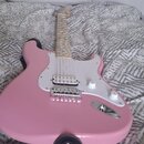 Růžová elektrická kytara s bílým pickguardem a světle hnědým hmatníkem. Nástroj má tři bílé snímače typu single-coil, dva bílé oválné knofíky a chrom…