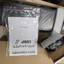Fotografie ukazuje otevřenou kartonovou krabici s polystyrenovými výplněmi, ve které je umístěn černý předmět v plastové fólii. V popředí je průhledn…