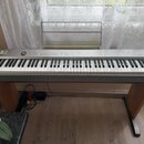 Digitální klavír v šedé barvě s dřevěným stojanem a notovým pulpitem. Na levé straně ovládacího panelu je logo ROLAND a vpravo nápis PS-20. Nástroj m…