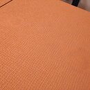 Detailní záběr na povrch oranžového pouzdra nebo kufru. Materiál má výraznou texturovanou strukturu připomínající tkaninu. Na povrchu nejsou vidět žá…