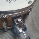 Detailní pohled na snare drum s bílým blátcem značky REMO, na kterém je nápis AMBASSADOR COATED a Made in Taiwan. Na blátci je vidět černý podpis. V…