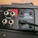 Detail zadní strany černého elektronického zařízení s konektory. Jsou zde čtyři jackové vstupy označené nápisy OUTPUT A, OUTPUT B a RETURN A, RETURN…