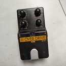 Černý efekt pedál s nápisem OVER DRIVE na žlutém pozadí a označením PEARL OD-05. Zařízení má čtyři ovládací knoflíky s popisky EQ GAIN, FREQUENCY, OU…