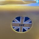 Detail žlutého plastového kufru nebo obalu. V kulatém výřezu je vidět nálepka s motivem britské vlajky a modrým nápisem 10T. Povrch materiálu vykazuj…