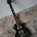 Černá elektrická kytara s tvarem typu single-cut a bílým lemováním. Na hlavě je bílé logo "Epiphone" a nápis "M.C. Cameron". Hmatník obsahuje vložky…