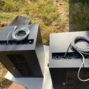 Dvě černé, kostkovité zařízení s kovovými mřížkami na přední straně. Na horní části obou přístrojů leží svitek šedého kabelu. Povrch zařízení vykazuj…