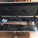Černá elektrická kytara v otevřeném tvrdém kufru s černým polstrováním. Na hlavě nástroje je bílé logo Epiphone a na hmatníku nápis Epiphone. Kytara…
