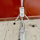 Stříbrný kovový stojan na hi-hat s trojnohým základnám a pedálem. Na bílém štítku je nápis HI-HAT STAND, MODEL NO H-830 a čárový kód. Na spodní části…
