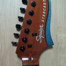 Hlava elektrické kytary z hnědého dřeva s modrým povrchem na hraně. Na hlave jsou vidět nápisy Squier BY FENDER a STARCASTER. Nástroj má šest černých…