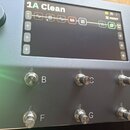 Detailní pohled na digitální procesor s displejem a ovládacími prvky. Na horní části je nápis QUAD CORTEX a displej s textem 1A Clean a zobrazením ef…