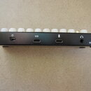 Detail zadní strany černého elektronického zařízení s kovovými konektory. Jsou zde viditelné nápisy IN a OUT u velkých jackových vstupů, nápis USB u…