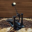 Fotografie znázorňuje pedál basového bubnu v černé barvě s kovovými mechanismy. Pedál je vybaven řetězovým přenosem a šlapkou s texturovaným povrchem…