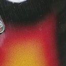 Detail korpusu elektrické kytary s barevným přechodem od černé přes červenou po žlutou. Povrch lakovaného dřeva vykazuje četné škrábance a drobné odr…