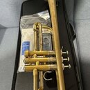 Zlatavý mosazný trumpet položený na černém kufru. Na horním ventilovém bloku je vyryto logo "CONN". Vedle nástroje se v plastovém obalu nachází modrá…