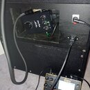 Fotografie zachycuje zadní část černého kytarového komba s vyřezem v korpusu, ve kterém je umístěno černé zařízení s nápisy „HardTube“ a „GT-100“. Po…