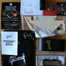 Sada příslušenství k hudebnímu nástroji rozložená na dřevěném povrchu. Je zde černý obal a černý kožený řemen s nápisem Gibson, bílá plastová krytka…