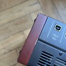 Detailní pohled na část elektronického zařízení s černým kovovým šasi a hnědým bočním panelem. Na černé části jsou vidět ventilace, USB port s nápise…