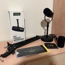 Na fotografii je sada mikrofonu a příslušenství položená na světlém dřevěném povrchu. Je zde bílá krabice s nápisy SHURE a MV7 PODCAST MICROPHONE, če…