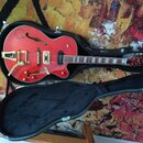 Červená polodupová elektrická kytara v černém tvrdém kufru. Na horní dece je vidět nápis "Epiphone" na hlavě hmatníku a zlatý hardware. Povrch nástro…