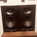 Dva černé studiové monitory v obdélníkovém provedení, umístěné vedle sebe na dřevěném stolku. Na spodní hraně obou reproduktorů je bílé logo a nápis…