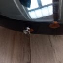 Detail spodní části černého, lesklého hudebního nástroje. Na hraně je vidět kovový konektor pro kabel a dvě nalepené hnědé plstěné podložky. Povrch v…