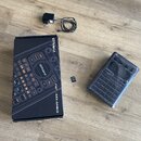 Na fotografii je sampler Roland SP-404MKII v průhledném plastovém obalu, černá krabice s nápisy Roland a SP-404MKII, napájecí adaptér s kabelem a mal…