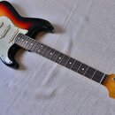 Elektrická kytara s trupem v barevném přechodu od žluté přes červenou k černé a bílým pickguardem. Na hlavě hmatníku je vidět nápis Fender STRATOCAST…