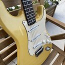 Žlutá elektrická kytara typu Stratocaster s bílým pickguardem a třemi snímači. Na povrchu těla je v horní části viditelné odřápání laku. Nástroj nemá…