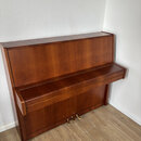 Hnědé dřevěné piano s uzavřenou klaviatuрой. Nástroj stojí v rohu místnosti na světlé dřevěné podlaze. V dolní části jsou vidět tři mosazné pedály. N…