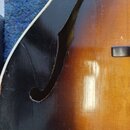 Detailní pohled na tělo kytary s f-otvorem. Povrch má tmavě hnědý až černý přechod do světlejší žlutohnědé barvy. Lak vykazuje drobné škrábance a opo…