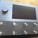 Stříbrný elektronický přístroj s matným povrchem. Na předním panelu je velký černý displej, vlevo ovládací knoflík s nápisem VOLUME a pod ním symbol…