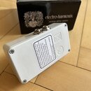 Bílý efekt pedál v kovovém krytu s dvěma konektory na horní straně. Na povrchu je bílá nálepka s anglickým textem začínajícím slovy „This device comp…