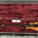 Elektrická kytara s korpusem v barvě sunburst a hnědým hmatníkem leží v otevřeném tvrdém kufru s červeným polstrováním. Na hlavě kytary je bílý nápis…