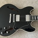 Černá polodupatá elektrická kytara s bílým lemováním a hnědým dřevěným vnitřkem ozvučnice. Nástroj má dva humbuckery, stříbrný most a čtyři černé ovl…